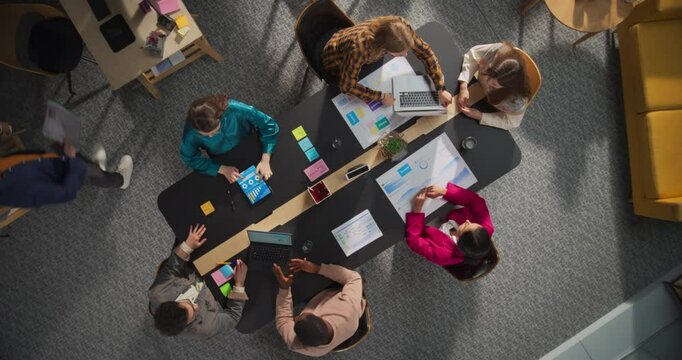 Top Down View Of Corporate Office Conference Room: Diverse Colleagues Gathered Around Table, Brainstorming Ideas, Business Plan, Marketing Strategy, Using Laptop And Tablet Computer, Discussing Charts