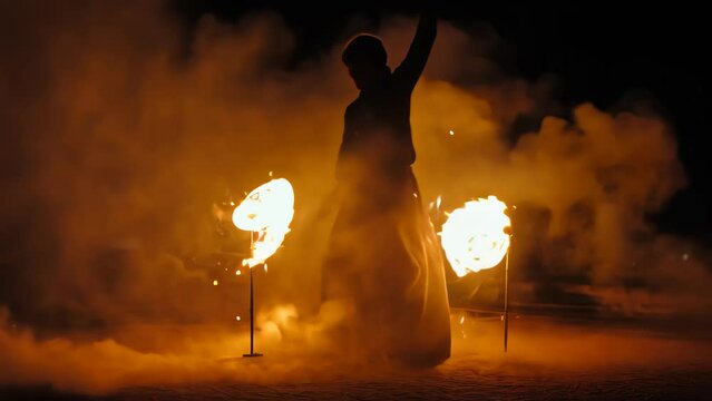 Skilled performer twirls flaming batons, creating mesmerizing circles of fire against dark backdrop. Intense orange glow illuminates artist's silhouette.