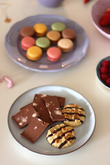 Plate of pastel macarons, cookies and chocolate, cup of tea of coffee, glass of bubble water, various berries, books and accessories on the table. Selective focus, pastel colors.