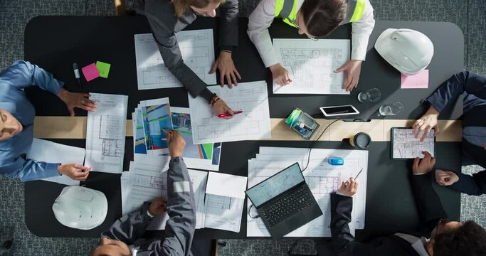 Top Down View of Diverse Team of Business Professionals and Construction Engineers Discussing Blueprints and Architectural Plans For Real Estate Projects in an Office. Teamwork With Tablet And Laptop.