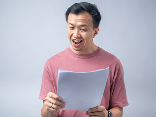 A man in a pink shirt looks excitedly at a stack of papers he is holding, his mouth open in...