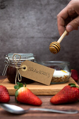 Delicious snack with cheesecake and homemade strawberry jam, glass jar with granola, yogurt and strawberry jam. Background with man pouring honey, vertical image for social media