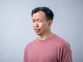 An Asian man in a pink shirt makes a skeptical and uncomfortable expression, as if reacting to something unpleasant. The plain light blue background highlights his displeased
