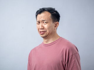An Asian man in a pink shirt makes a skeptical and uncomfortable expression, as if reacting to something unpleasant. The plain light blue background highlights his displeased