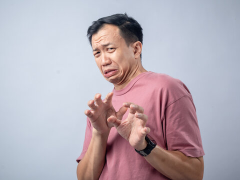 A man in a pink shirt makes a disgusted and fearful expression with his hands raised in a defensive gesture. The plain light blue background emphasizes his reaction