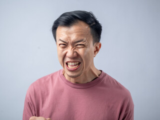 An Asian man in a pink shirt exhibits a fierce expression, baring his teeth and raising his hands with claw-like gestures, as if in anger or frustration. The plain light blue background highlights