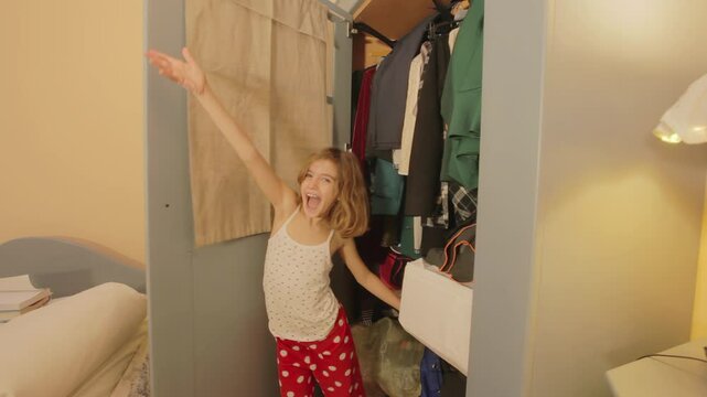 A cute little girl emerges from the wardrobe in her bedroom, smiles, and then returns inside after making a shushing gesture. Long shot.
