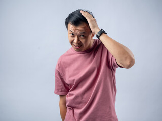 A cheerful Asian man in a pink t-shirt smiles while scratching his head with one hand. The studio background is light grey, emphasizing his casual attire and relaxed demeanor