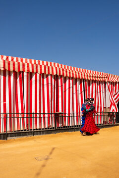 A vibrant scene at a traditional fair with a red and white striped tent as the backdrop, and people dressed in colorful, festive attire walking along the sandy path in Seville in Spain