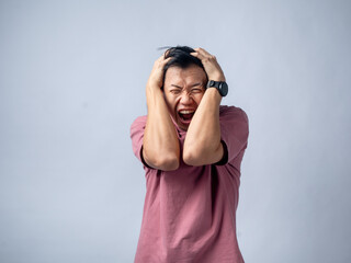 Asian man in a pink t-shirt with a frustrated expression, pulling his hair with both hands, standing against a plain background. The image captures a moment of intense stress or anger