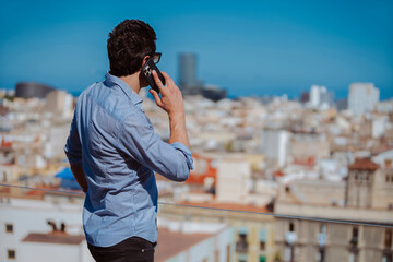 Rear view of businessman talking with smartphone on balcony outdoors. Casual strong man working in front of city skyline during a phone call.