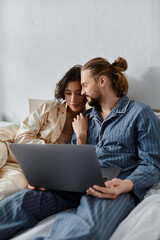 A loving couple cuddles in bed while browsing on a laptop.