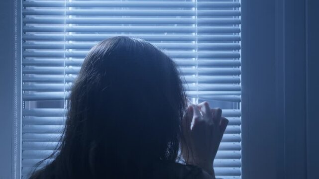 A frightened woman looks out of the window through the sting from inside the house. Blue light falls on her from the window