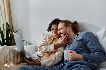 A couple cuddles in bed, enjoying a morning with coffee and a laptop.