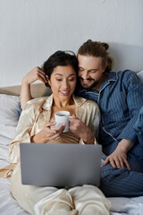 A loving couple relaxes in bed, enjoying coffee and a laptop.