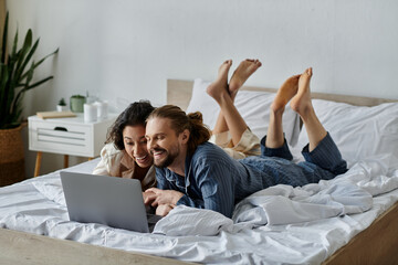 A loving couple cuddles in bed, sharing a laugh while browsing the internet on a laptop.