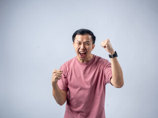 A man in a pink shirt raises both fists in a celebratory gesture, his face beaming with excitement and joy. The plain light blue background emphasizes his triumphant and enthusiastic expression.