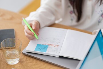 Close-up of a person writing notes in a notebook at a desk with a green pen A glass and a smartphone are also on the desk