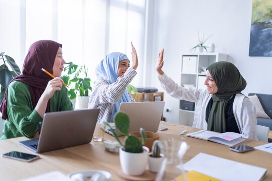 Three women in hijabs giving each other high-fives at their office desk