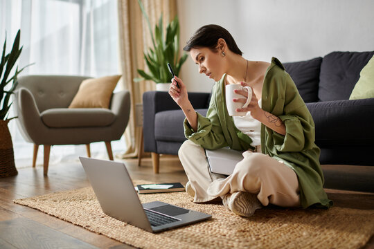 A woman sits on a rug in her home office, working on her laptop with a cup of coffee in hand.