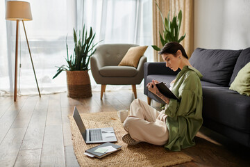 A woman sits on the floor in a home office, working on a laptop and taking notes.