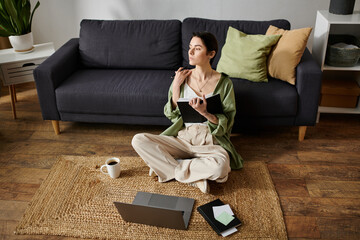 A woman sits on a rug in her living room, working on a laptop and taking notes in a notebook.