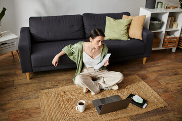 A woman sits on a rug in her home, working on a laptop.
