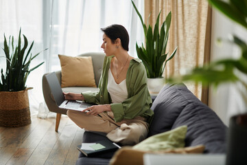 An attractive woman works remotely from home, comfortably seated on a sofa in a well-lit room.
