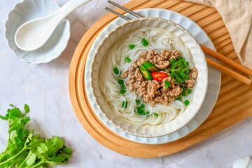 Rice vermcelli  clear soup, instant noodles clear soup toped with minced pork and chili and vegetables in a white bowl with chopsticks and white spoon, top view food table
