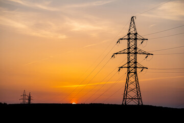 High voltage pylons and power lines against orange sunrise near woodland