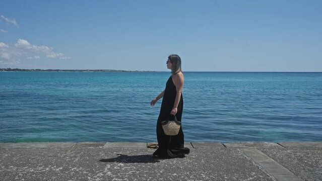 Young woman on a sunny day in porto cesareo, salento, walking by the blue water, wearing sunglasses and a black dress, holding a straw bag.