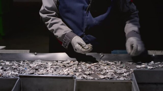 Worker sorts trash on conveyor belt at waste recycling plant. Metal pieces falling into a box from recycling processing machine. Man sorts pieces of metal, iron or steel with his hands