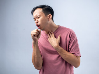 A man in a pink shirt coughs violently, holding his throat with one hand and covering his mouth with the other. His expression shows discomfort and pain. The plain light blue background