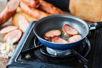 Step-by-step cooking of the dish. Pieces of sausage are fried in a frying pan on a camp stove.