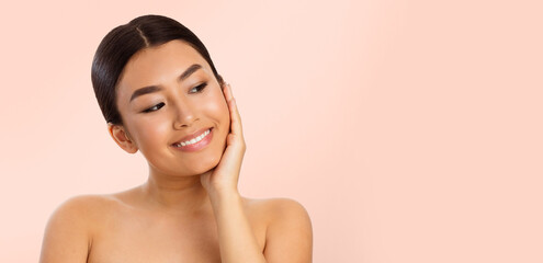 A young Asian woman with dark hair pulled back smiles and looks at copy space while touching her face. She is wearing no makeup and has a soft, natural look. The background is a light pink color.