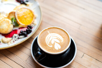 Top view of cappuccino with latte art and cottage cheese pancakes on wooden table in cozy coffee shop. Close-up food and drink breakfast scene.