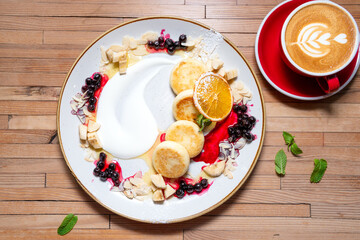Top-down shot of homemade pancakes with berry jam on white plate and cappuccino on wooden table. Cozy breakfast setup, flat lay composition