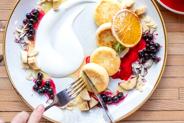 Top view of a woman eating syrniki — cottage cheese fritters with strawberry jam. Female hands holding fork and knife over homemade breakfast pancakes.	