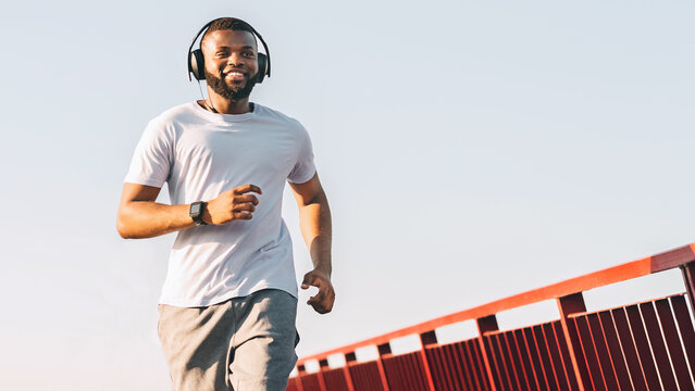 A man in a white t-shirt and headphones is running on a bridge with a red railing. He is smiling and looks happy.