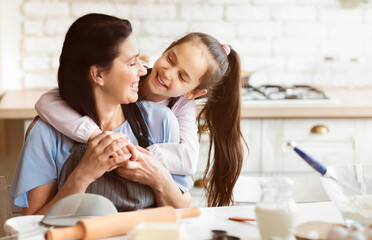 A young girl is hugging her mother from behind while they are in the kitchen, likely baking together. They are both smiling and happy, enjoying each others company, copy space