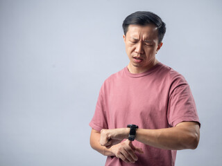 An Asian man in a pink shirt looks at his watch with a concerned and impatient expression,...