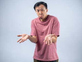 Naklejka premium An Asian man in a pink shirt shrugs with a confused and uncertain expression, his arms outstretched and palms up. The plain light blue background emphasizes his ambiguous
