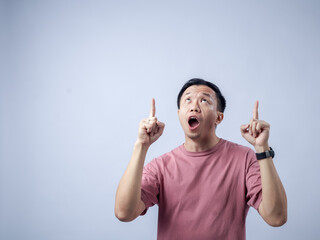 Asian man in a pink t-shirt, standing against a plain background, pointing upwards with both index...