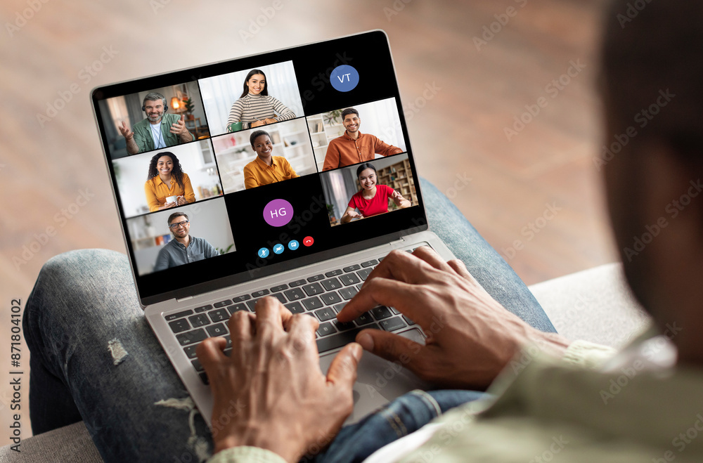 Aufkleber Black man sits on a couch, using a laptop computer to participate in a video conference meeting with several other people. The laptop screen is displaying a grid of video feeds
