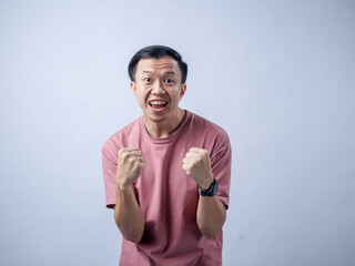 Fototapeta premium A man in a pink shirt raises both fists in a celebratory gesture, his face beaming with excitement and joy. The plain light blue background emphasizes his triumphant and enthusiastic expression.