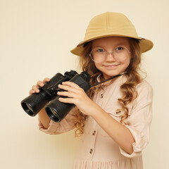 Traveler child. Child discoverer. Young researcher. Little girl in a safari hat with binoculars in her hands. Concept: search for adventure and treasure. Cute happy girl in safari clothes