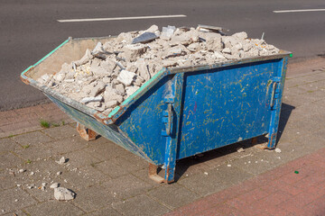 Kleine blaue Absetzmulde oder Müllcontainer mit Bauschutt gefüllt auf der Straße stehend,...
