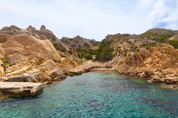 Fototapeta premium Cala Candeo en la isla de Caprera, parte del archipiélago de la Magdalena, Cerdeña, Italia. Una pequeña cala rodeada de formaciones rocosas de tonos anaranjados y vegetación mediterránea.