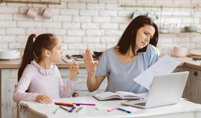 A busy mother works from home while her young daughter draws at the kitchen table. The mother is on...