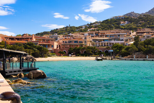 Vista de Porto Cervo en Cerde&ntilde;a, Italia, en un d&iacute;a soleado. Las aguas turquesas del mar rodean un muelle y una playa de arena, con villas de color terracota construidas en la ladera. 